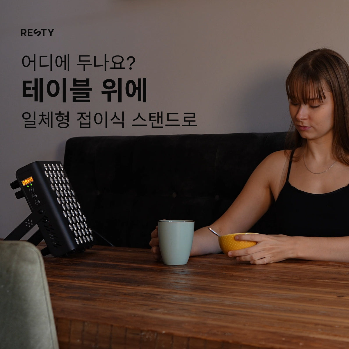 한 여성이 머그잔과 그릇이 놓인 나무 식탁에 앉아 있습니다. 그 옆 식탁 위에는 MARS 400이 일체형 접이식 탁상용 스탠드로 세워져 있으며, 별도의 바닥 스탠드는 없습니다.